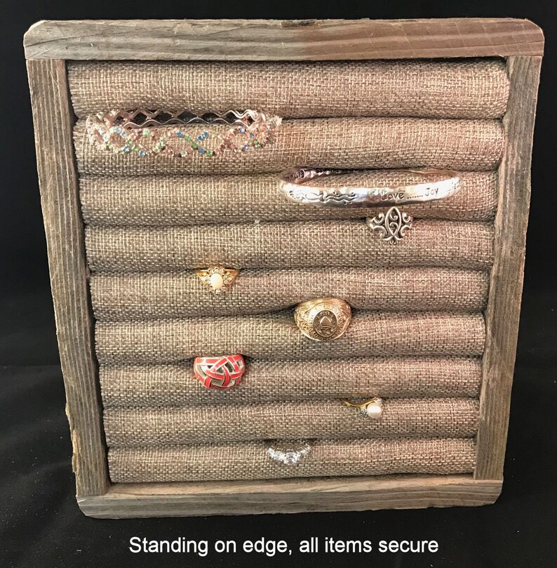 May include: A wooden ring holder with a burlap fabric interior. The holder has multiple slots for rings and other jewelry. The holder is standing on its edge, and all of the items are secure.