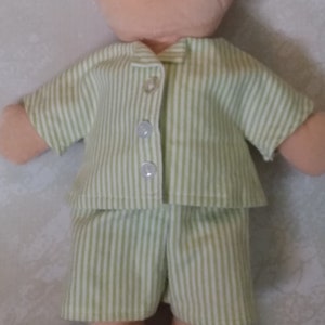 Può includere: Una bambola che indossa un pigiama a righe verdi e bianche. La camicia a maniche corte ha tre bottoni trasparenti. I pantaloncini abbinati completano l'outfit. Il viso della bambola è visibile nella parte superiore dell'immagine.