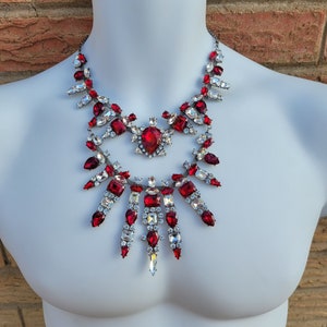 May include: A statement necklace with a large red and clear crystal design. The necklace is made of metal and features a variety of crystal shapes and sizes.