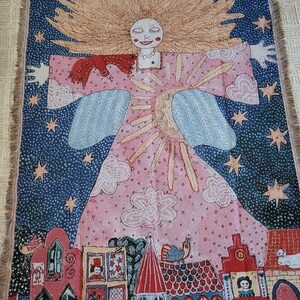 Op de afbeelding: Een geweven sprei met een grillig ontwerp. Het kunstwerk toont een lachende figuur met vloeiend blond haar, een rode vogel en een dorpssc&egrave;ne. De achtergrond is een sterrenhemel met roze en blauwe accenten.