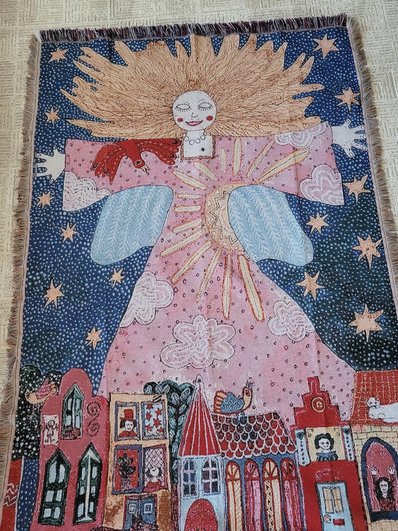 Op de afbeelding: Een geweven sprei met een grillig ontwerp. Het kunstwerk toont een lachende figuur met vloeiend blond haar, een rode vogel en een dorpssc&egrave;ne. De achtergrond is een sterrenhemel met roze en blauwe accenten.