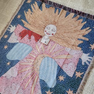 Op de afbeelding: Een geweven sierdeken met een grillig ontwerp. Het toont een lachende figuur met lang blond haar, omringd door een zonnestraal, sterren en wolken. De achtergrond is roze, blauw en wit, met franjes.