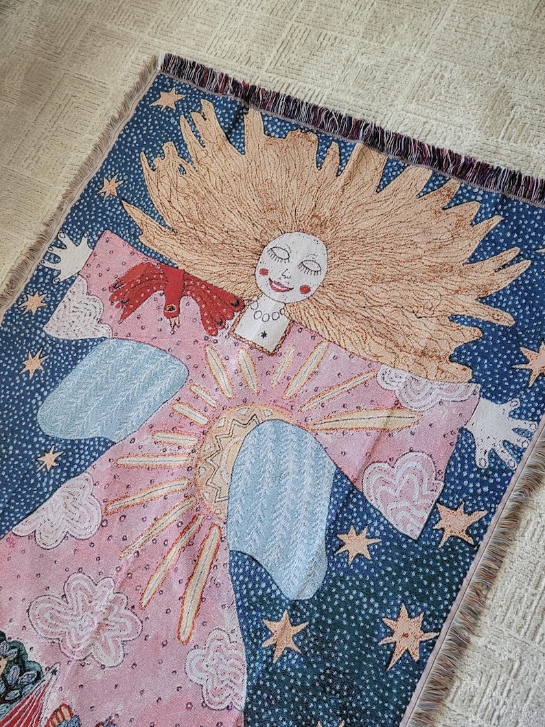 Op de afbeelding: Een geweven sierdeken met een grillig ontwerp. Het toont een lachende figuur met lang blond haar, omringd door een zonnestraal, sterren en wolken. De achtergrond is roze, blauw en wit, met franjes.