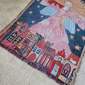 Op de afbeelding: Een geweven sprei met een grillige illustratie. Het ontwerp omvat een lachende zon, sterren, wolken en een dorp met rode en blauwe gebouwen. De sprei heeft een gefranjerde rand en is voornamelijk roze en blauw.