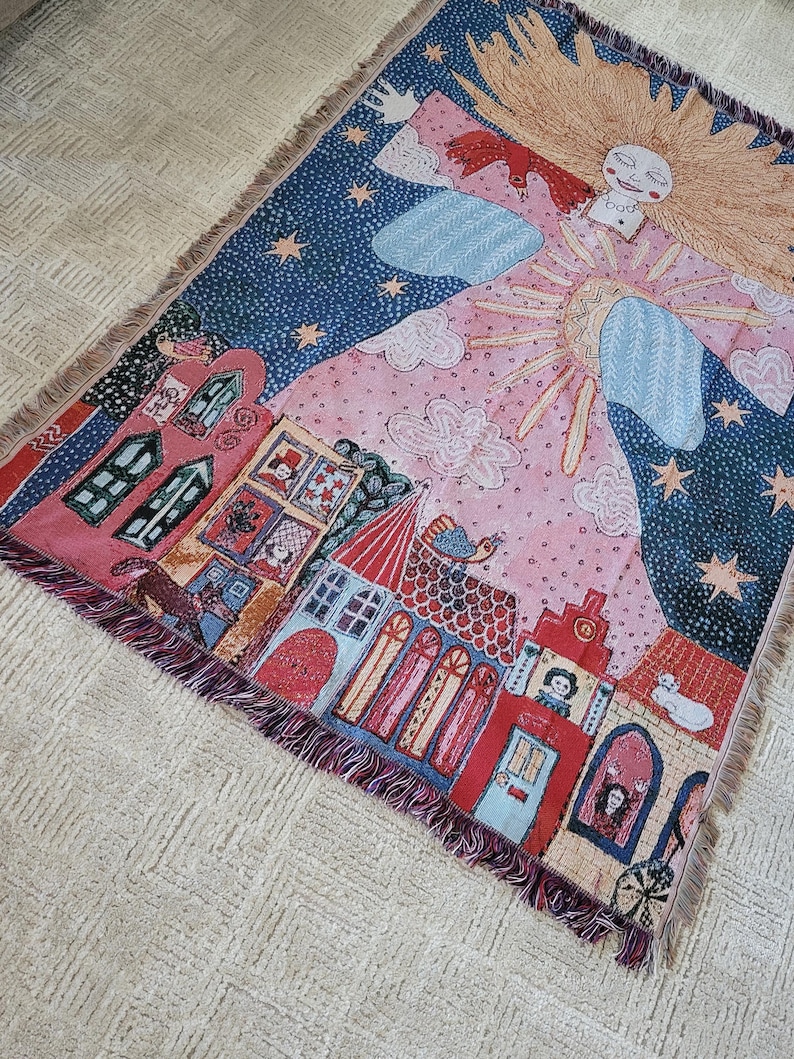 Op de afbeelding: Een geweven sprei met een grillige illustratie. Het ontwerp omvat een lachende zon, sterren, wolken en een dorp met rode en blauwe gebouwen. De sprei heeft een gefranjerde rand en is voornamelijk roze en blauw.