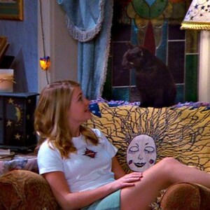 Op de afbeelding: Een sc&egrave;ne uit een televisieshow met een vrouw in een wit t-shirt en een zwarte kat. De vrouw zit op een bruine bank met een geel en blauw patroon. Een zwarte kast met sterren en manen is zichtbaar.
