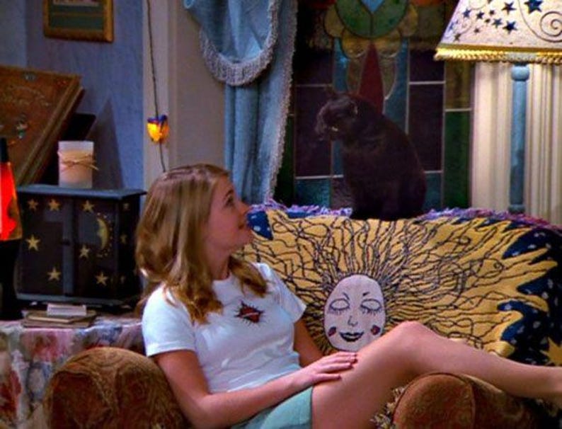 Op de afbeelding: Een sc&egrave;ne uit een televisieshow met een vrouw in een wit t-shirt en een zwarte kat. De vrouw zit op een bruine bank met een geel en blauw patroon. Een zwarte kast met sterren en manen is zichtbaar.