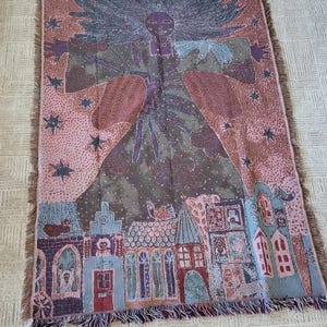 Op de afbeelding: Een geweven sprei met een hemels ontwerp. Het ontwerp toont een centrale figuur, sterren en een dorpssc&egrave;ne. Het kleurenpalet omvat tinten roze, paars, blauw en bruin. De deken heeft een gefranjerde rand.