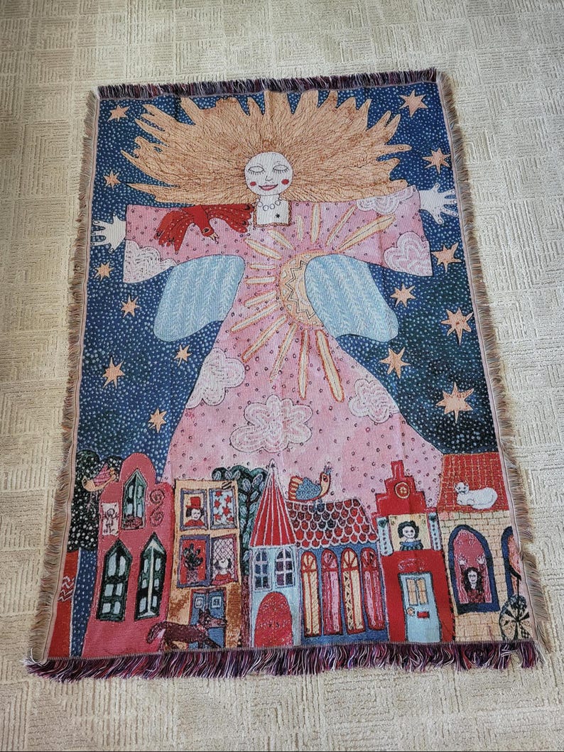 Op de afbeelding: Een geweven sprei met een grillig ontwerp. De sprei toont een lachende engelenfiguur met een sterrenhemel en een dorpssc&egrave;ne. Het kleurenpalet omvat roze, blauw en rood, met een gefranjerde rand.