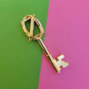 May include: A gold-colored key with a decorative top and a classic keyhole design. The key is set against a background of green and pink. The key's design includes a shield-like shape with a stylized letter or symbol.