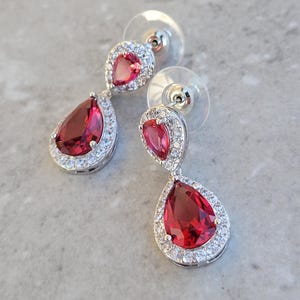 May include: A pair of silver dangle earrings with red pear-shaped gemstones surrounded by a halo of smaller clear gemstones.