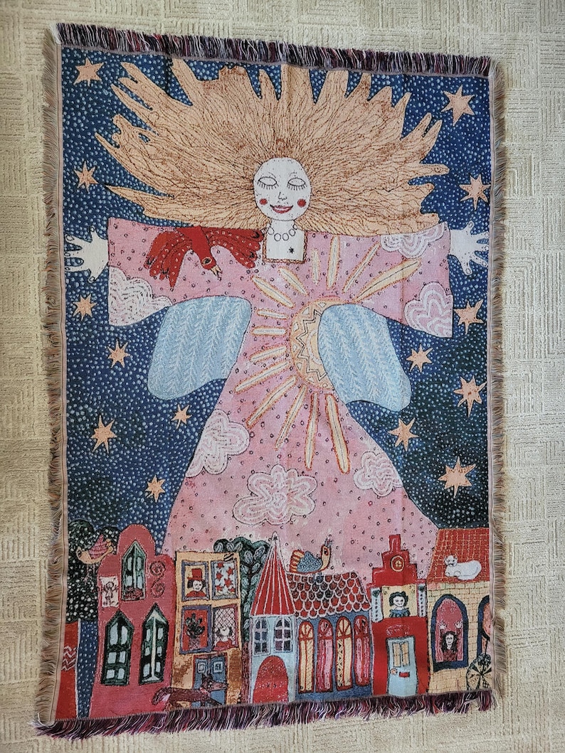 Op de afbeelding: Een geweven wandtapijt met een grillige illustratie. Het toont een figuur met golvend blond haar, een roze jurk en uitgestrekte armen. De achtergrond is een sterrenhemel met een rij kleurrijke huizen aan de onderkant. Het wandtapijt heeft een gefranjerde rand.