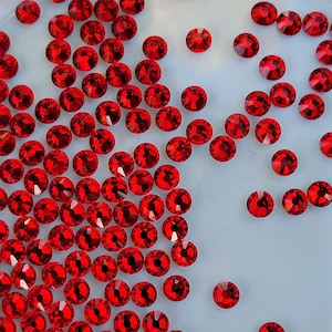 May include: A close-up image of a large quantity of small, red, round rhinestones. The rhinestones are scattered on a white surface.