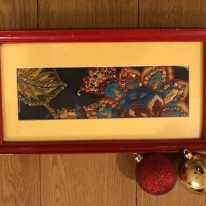 May include: A framed artwork with a colorful floral design in shades of blue, red, gold, and green. The frame is red with a yellow mat.