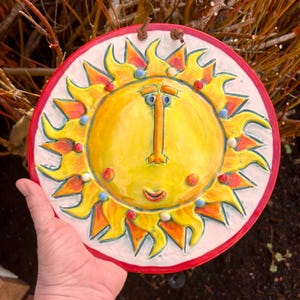 May include: A round, ceramic sun face wall hanging. The sun has a yellow center with orange and yellow rays, a red border, and a white background. The sun has a smiling face with blue and red accents. Two brown leather loops are at the top.