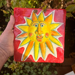 May include: A square, hand-painted ceramic sun face with a smiling expression. The sun has yellow and orange rays, a yellow face, and a red background. The artwork is held by a hand and has a leather cord for hanging.