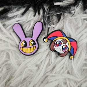 May include: Two decorative shoe charms. One charm is a purple rabbit head with long ears, a wide yellow grin, and cross-shaped eyes. The second charm is a character wearing a red and blue jester hat with bells, featuring large eyes with red and blue pupils.