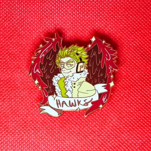 May include: A gold-colored enamel pin featuring a cartoon character with green hair and red wings. The character is wearing a white fur collar and a yellow jacket. The pin says "HAWKS" in a white banner.