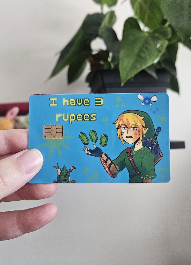 May include: A rectangular credit card skin featuring a light blue background with the text I have 3 rupees in yellow pixelated font. The design includes an illustration of a blonde character in a green tunic holding three green gems.