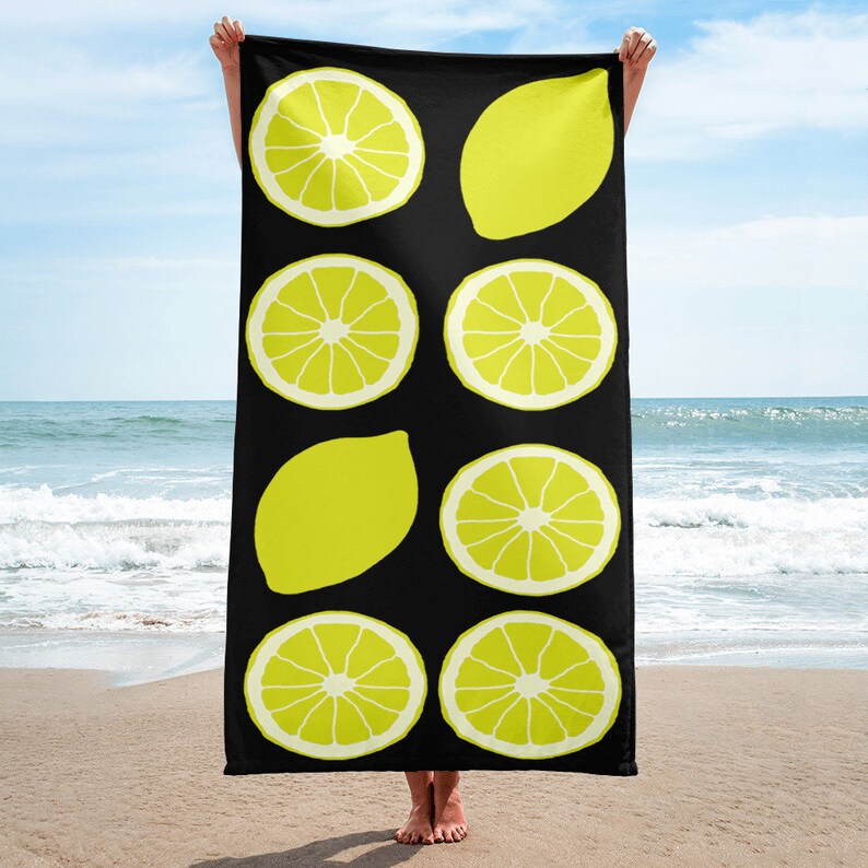 May include: A black beach towel with a repeating pattern of yellow lemons. The lemons are in various shapes and sizes, some are whole and some are sliced.