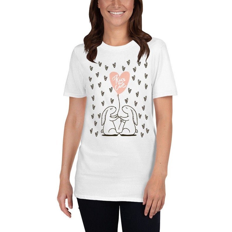 May include: White t-shirt featuring a graphic of two cartoon bunnies holding hands, with a pink heart-shaped balloon that says "Kiss Love". Small black hearts are scattered around the design.
