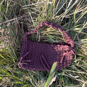 May include: A dark purple, hand-crocheted handbag with a braided handle and fringe details. The bag is rectangular with a rounded top and is lying in a bed of green and brown grass.