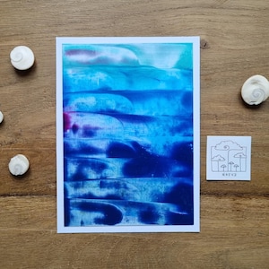 Könnte beinhalten: Ein abstrakter Kunstdruck mit Blau- und Weißtönen, der an Wasser oder Himmel erinnert, wird auf einer Holzoberfläche präsentiert. Kleine weiße Muscheln sind um den Druck herum verstreut. Ein kleiner Aufkleber mit einem Pilzdesign ist ebenfalls vorhanden.