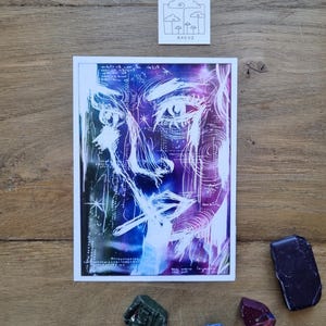 Può includere: Una stampa artistica di un volto con uno sfondo cosmico, con arte lineare bianca su un gradiente viola e blu. La stampa ha un bordo bianco. Sono presenti anche piccoli cristalli colorati e un piccolo adesivo quadrato con un disegno di nuvole e alberi.