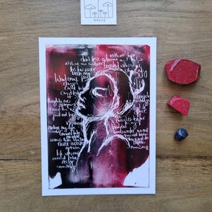 Può includere: Una stampa d'arte astratta con il profilo bianco di una donna su uno sfondo rosso e nero. Il testo scritto a mano circonda la figura. Una piccola carta con un disegno di nuvole e funghi è sopra la stampa.