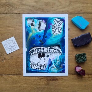 Può includere: Una stampa d'arte astratta con bordo bianco, con uno sfondo blu e verde acqua con elementi grafici in bianco e nero. È presente anche un piccolo adesivo con un disegno di nuvole e funghi, insieme a diverse piccole pietre.