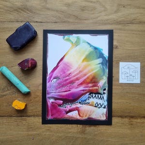 Può includere: Un'opera d'arte astratta in una cornice nera, con una vibrante miscela di rosa, giallo e blu. Accanto, materiali artistici: un pastello blu scuro, un pastello turchese e pezzi di pastelli rossi e arancioni. È presente anche un piccolo quadrato con un disegno di nuvole e alberi.