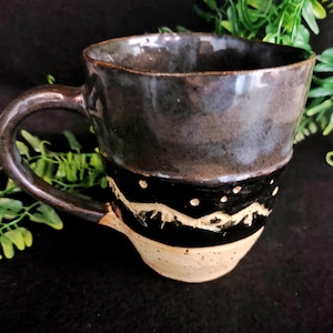 Op de afbeelding: Een handgemaakte keramische mok met een donkerbruin glazuur. De mok heeft een zwarte band met een wit bergontwerp en witte stippen. De onderkant van de mok is lichtbeige van kleur. De handgreep is donkerbruin.