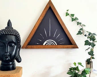 Puede incluir: Un marco de madera triangular con fondo negro y un diseño de sol blanco. El marco está colgado en una pared blanca.