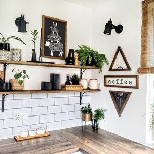 May include: A white kitchen with a wooden shelf and white tiled backsplash. The shelf is decorated with plants, jars, and a black and white print of a butcher's chart. There are also two black light fixtures on the wall. The text "coffee" is written on a wooden sign.