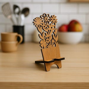 Könnte beinhalten: Ein Handyhalter aus Holz mit floralem Design. Der Ständer ist aus hellbraunem Holz gefertigt und hat einen Ausschnitt aus Blumen und Blättern. Die Basis hat zwei kleine Vorsprünge, um ein Telefon zu halten. Der Ständer steht auf einer hellbraunen Holzoberfläche.