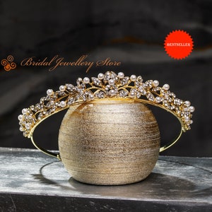 Op de afbeelding: Een goudkleurige tiara met een bloemmotief, versierd met heldere kristallen en imitatieparels. De tiara wordt gepresenteerd op een gouden decoratieve bol. De tekst "Bridal Jewellery Store" is zichtbaar. Een rode "Bestseller" sticker is ook zichtbaar.