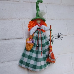 May include: A hanging doll ornament featuring a crocheted green witch hat, bright orange curly hair, and a white blouse. The doll wears a green and white checkered skirt, carries a small yellow crocheted basket, and holds an orange broom with a silver crescent moon charm.
