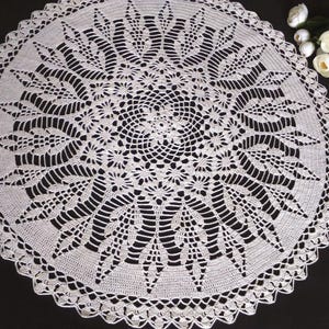 Könnte beinhalten: Weißes, rundes, gehäkeltes Deckchen mit einem komplizierten Blumen- und Blättermuster. Das Deckchen hat einen gewellten Rand und ein zentrales Design, das sich nach außen ausdehnt. Die detaillierte Lochmusterung erzeugt ein zartes und elegantes Aussehen, geeignet für die Heimdekoration.