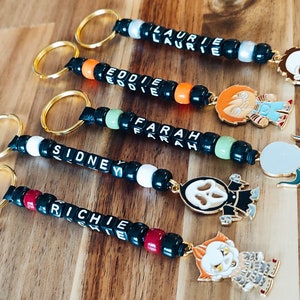 Puede incluir: Cinco llaveros de cuentas coloridas con cuentas negras y cuentas de letras blancas que deletrean nombres. Cada llavero tiene un amuleto de personaje diferente, incluyendo un fantasma, un payaso, un hombre con un cuchillo, un hombre con un gancho y un hombre con una máscara.