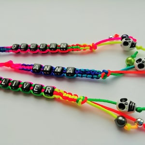 Peut inclure: Trois porte-clés colorés avec un cordon arc-en-ciel et des perles de lettres noires orthographiant les noms "Declan", "Anna" et "Oliver". Chaque porte-clés a une perle de crâne blanche avec des yeux noirs et une perle argentée.