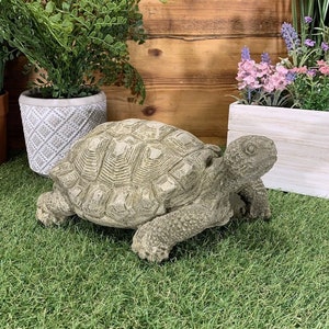 May include: A grey concrete garden statue of a turtle with its head raised. The turtle is sitting on a green artificial grass surface.