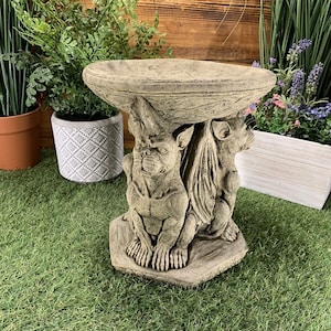 May include: A grey stone bird bath with a bowl shaped like a bird's nest. The base of the bird bath is sculpted with two gargoyles.