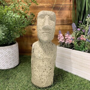May include: A grey stone statue of a human head with a large nose and closed eyes. The statue is standing on a green grassy surface.