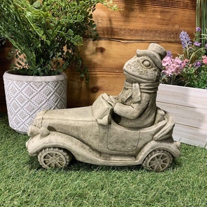 May include: A grey stone sculpture of a frog wearing a hat and scarf driving a vintage car. The sculpture is on a green artificial grass surface.