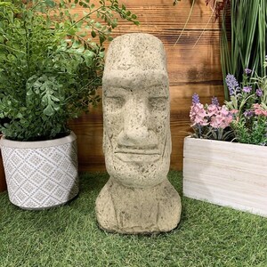 May include: A weathered stone Moai head statue with carved facial features. The sculpture is a light beige colour and sits on a green surface, with potted plants in the background. The statue has a textured surface and a flat base.