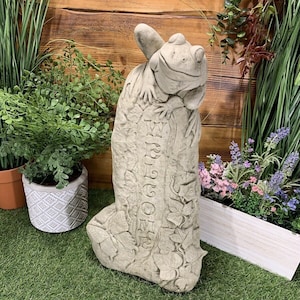 May include: A grey stone frog sculpture with a welcome sign on its back. The frog is sitting on a stone with leaves and vines carved into it. The sculpture is on a green grassy surface.