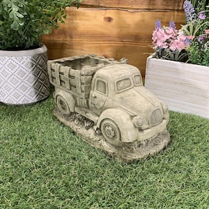 May include: A grey concrete planter shaped like a vintage pickup truck with a wooden bed. The truck is sitting on a green artificial grass surface.