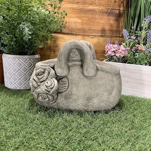 May include: A grey concrete garden sculpture shaped like a handbag with a floral design. The handbag has a handle and a floral design on the side.