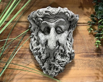 Op de afbeelding: Een grijs stenen beeld van een gezicht met een lange baard en een ondeugende uitdrukking. Het beeld is bevestigd aan een houten muur.
