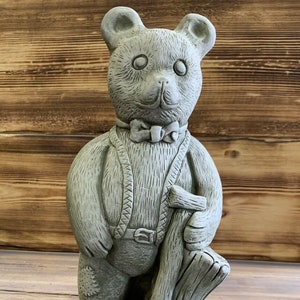 May include: A grey stone garden statue of a teddy bear wearing overalls and a bow tie, holding a shovel and standing on a base with leaves.